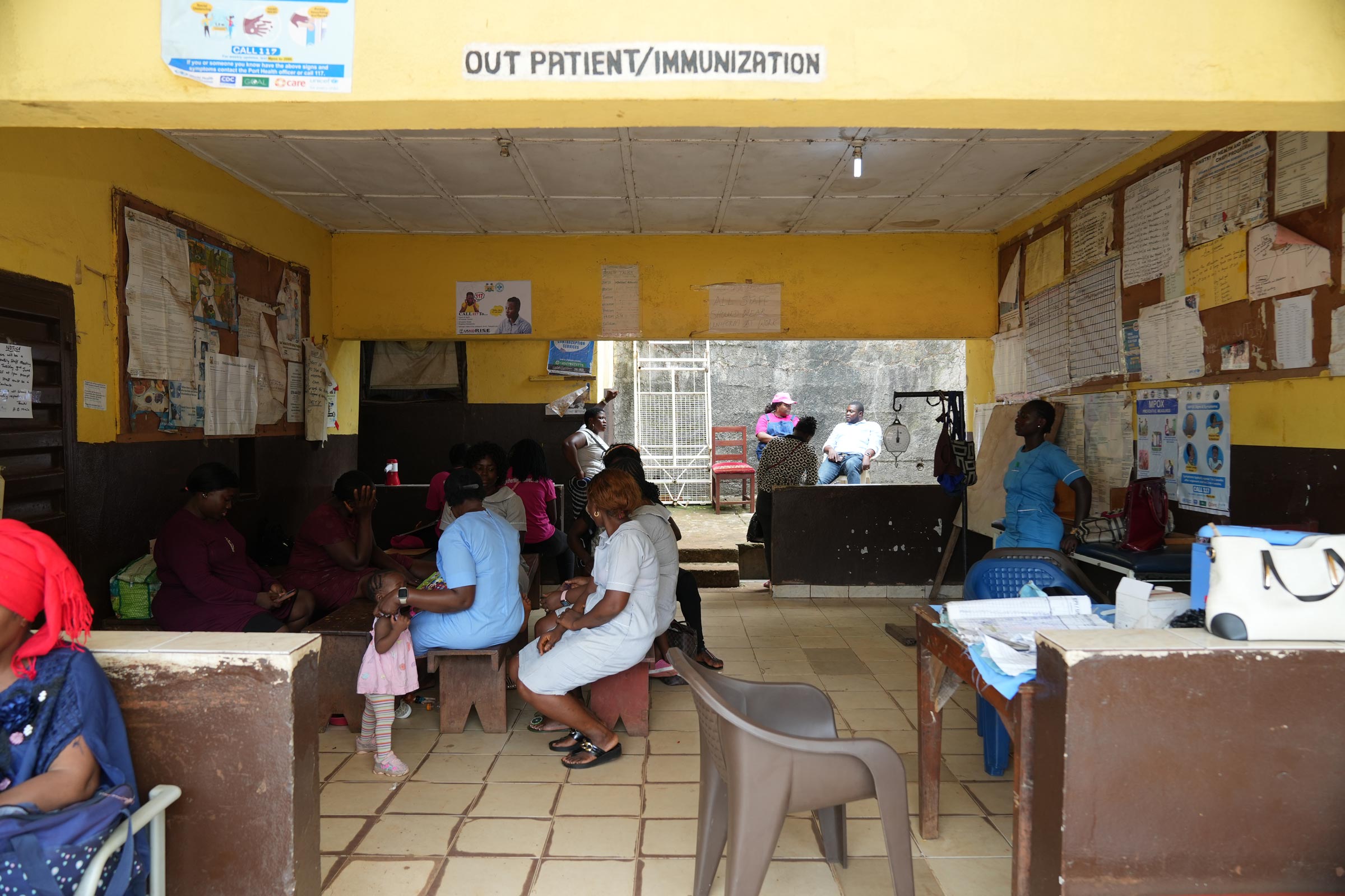 A community outpatient and immunization clinic with yellow and brown walls, where several women and children are seated on wooden benches