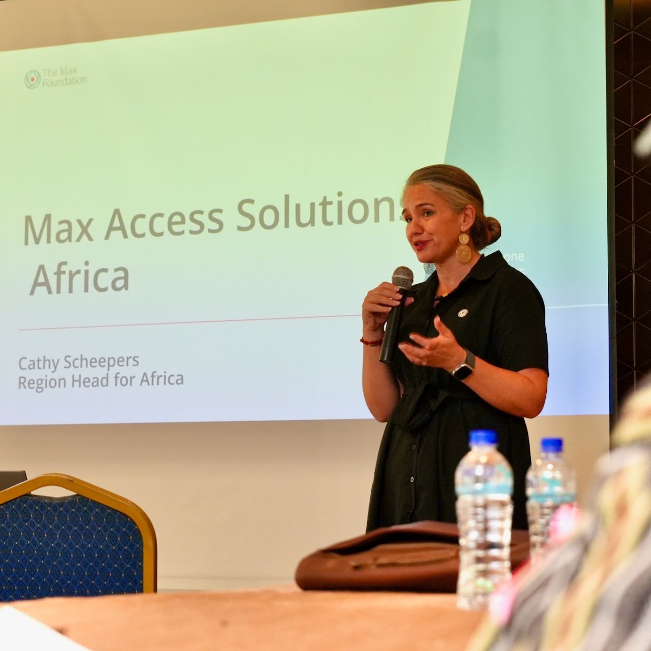 Cancer Programs, Cathy stands and speaks into a microphone in front of a presentation slide titled “Max Access Solutions Africa.”