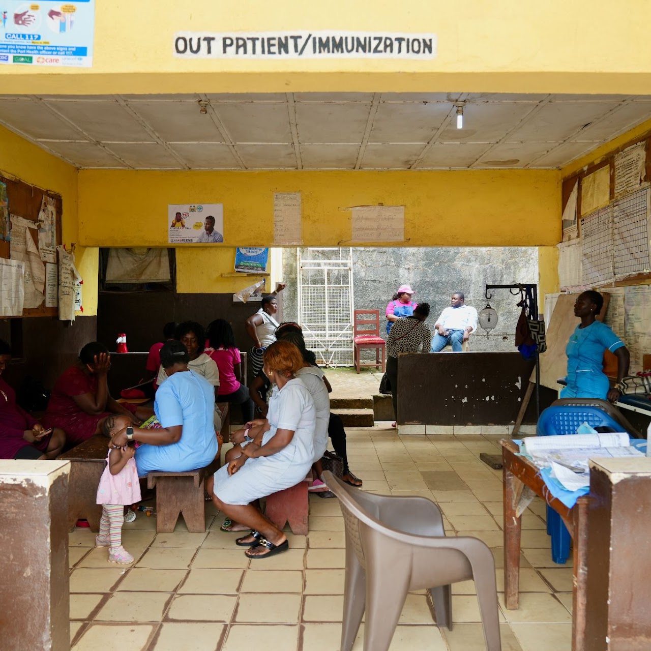 Our Work, A small clinic area labeled “Out Patient/Immunization”. Several people, including women and a child, are seated on wooden benches waiting.