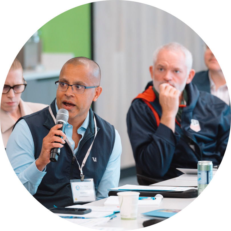 Participate with The Max Foundation. A person with a microphone sitting at a meeting table with others.
